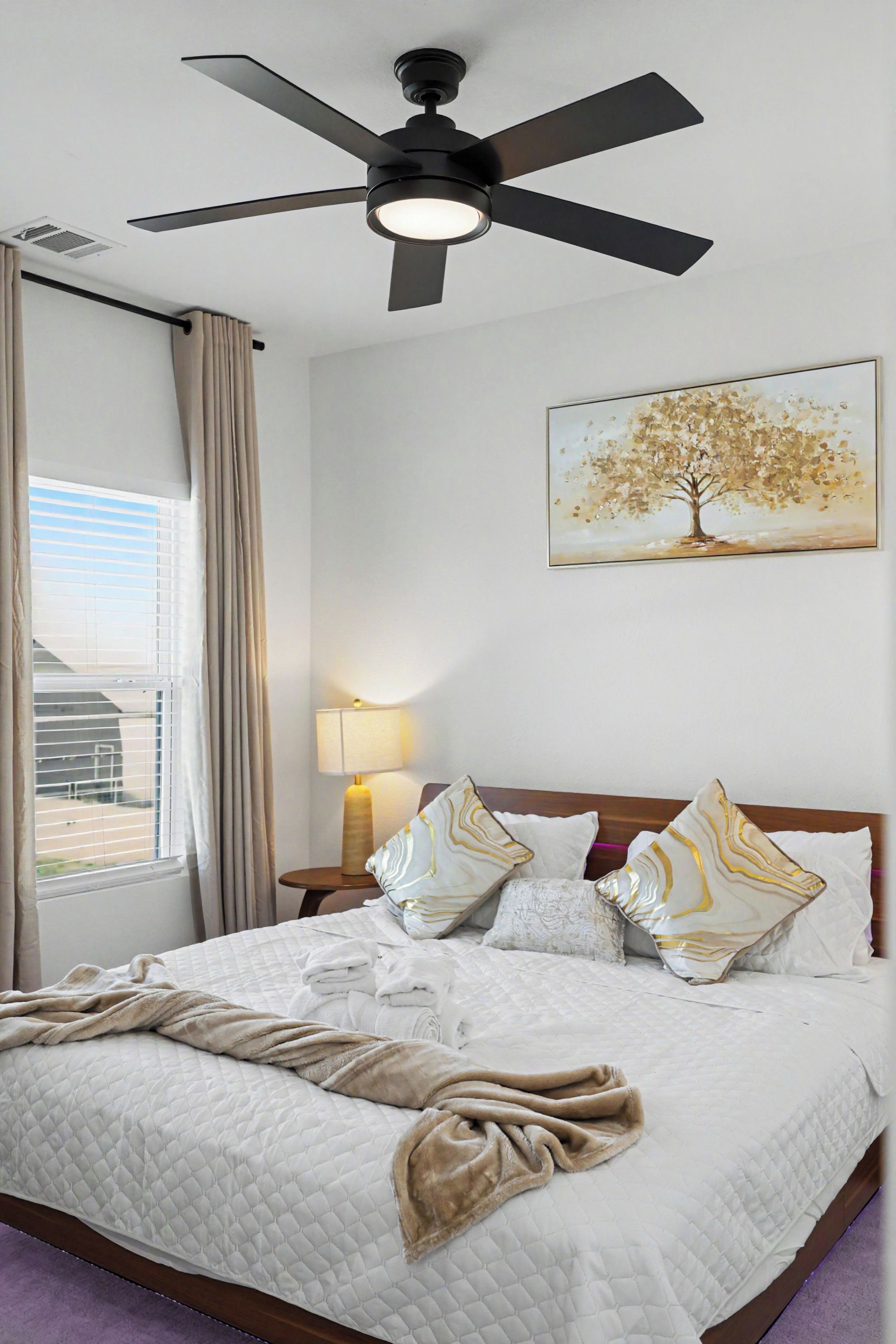 Une chambre d’hôtel avec un lit, un ventilateur de plafond, une fenêtre avec des rideaux, une lampe de chevet et un tableau au mur.