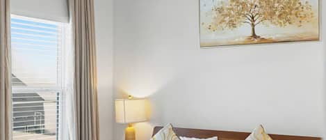 Une chambre d’hôtel avec un lit, un ventilateur de plafond, une fenêtre avec des rideaux, une lampe de chevet et un tableau au mur.