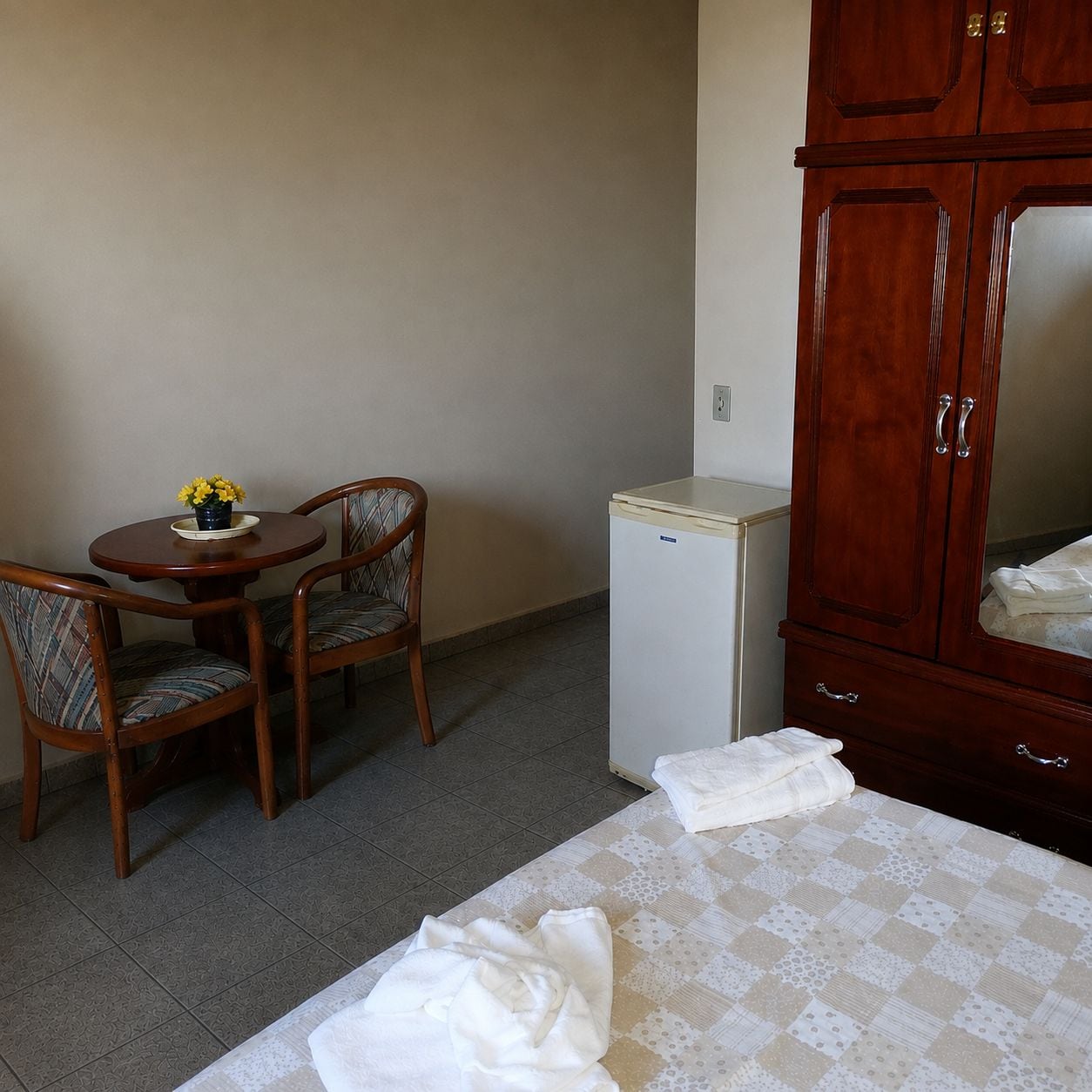 Una stanza con un letto, un piccolo frigorifero, un armadietto in legno, un tavolo rotondo con due sedie e un vaso con fiori gialli.