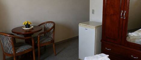A room with a bed, a small refrigerator, a wooden cabinet, a round table with two chairs, and a vase with yellow flowers.