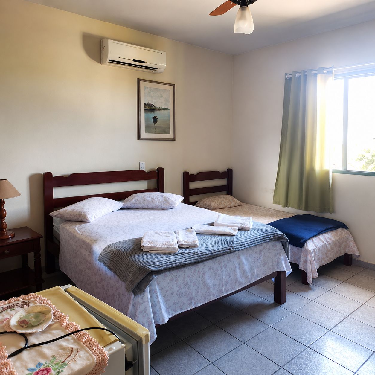 A hotel room with two beds, an air conditioning unit, a window with curtains, and a bedside table with a lamp.