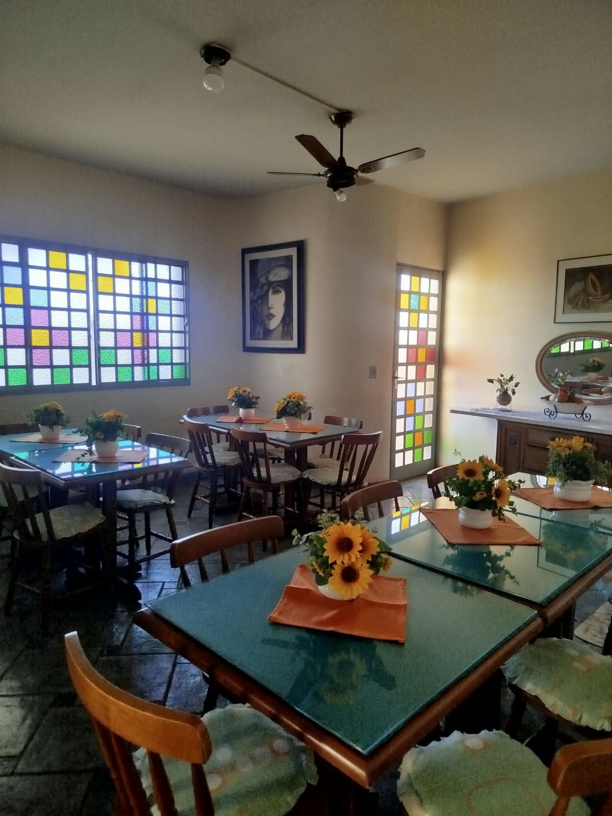 A dining area with glass tables and chairs, a ceiling fan, and a large window with colorful stained glass.