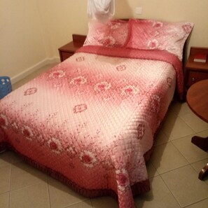 A bed with a patterned bedspread and pillows, a wooden nightstand, and a blue chair. - Blue Bay Hotel (Nakuru)