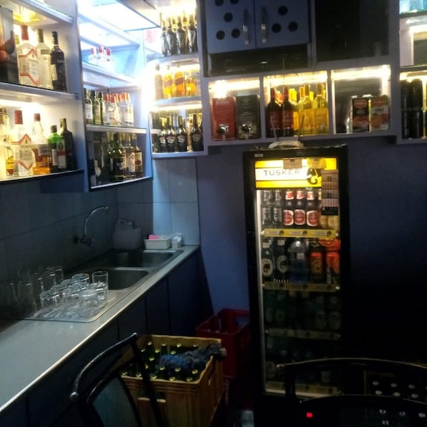 A bar area with shelves of liquor bottles, a refrigerator with beverages, and a counter with glasses. - Blue Bay Hotel (Nakuru)