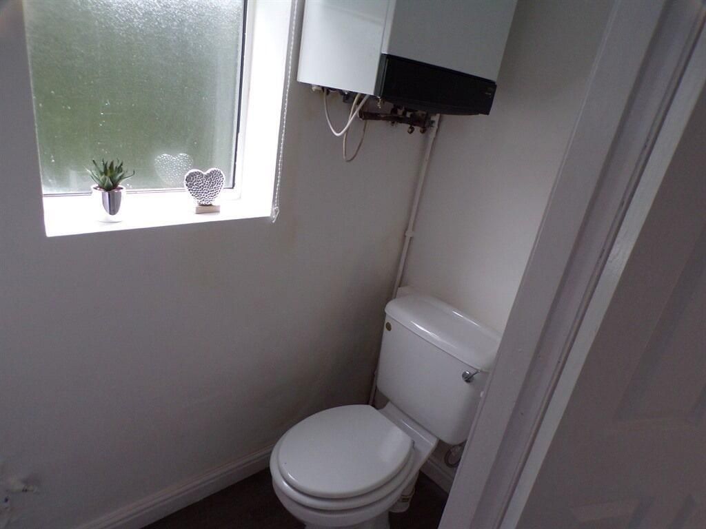 A small, compact bathroom with a toilet, a window with a plant and a heart-shaped decoration, and a white cabinet above the toilet.