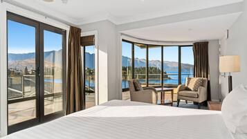 A hotel room with a large bed, two armchairs, a small table, and a view of mountains and a lake.
