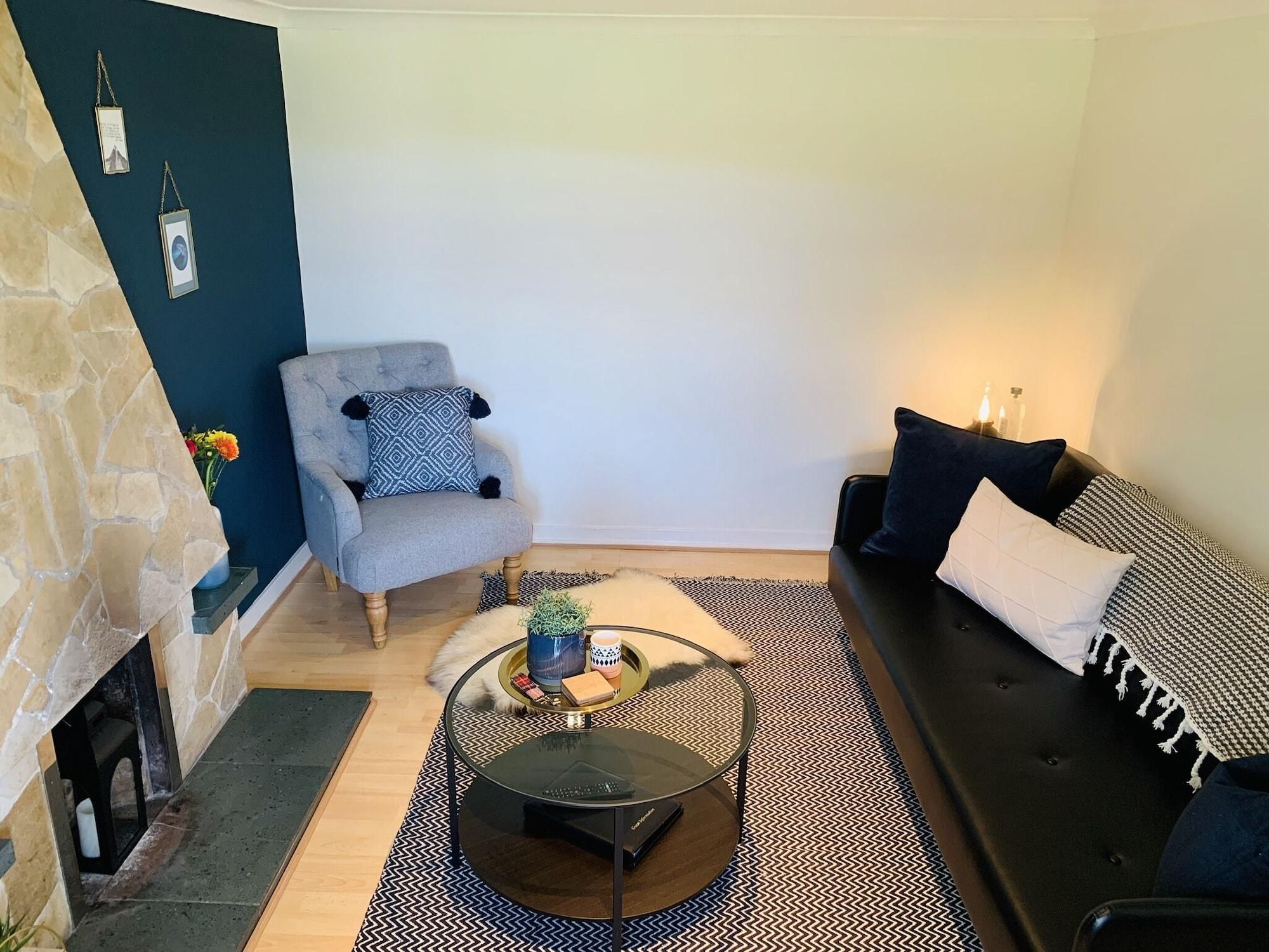 Un salon doté d’une cheminée en pierre, d’un fauteuil gris agrémenté d’un coussin à motifs, d’un canapé noir avec des coussins blancs et à motifs, et d’une table basse ronde avec un plateau en verre.