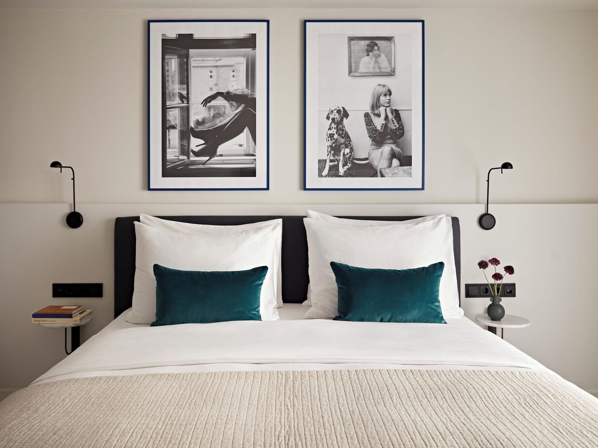 A neatly made bed with white and teal pillows, two framed black and white photographs on the wall, a bedside table with a book and a vase with flowers, and a modern lamp.