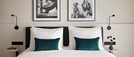A neatly made bed with white and teal pillows, two framed black and white photographs on the wall, a bedside table with a book and a vase with flowers, and a modern lamp.