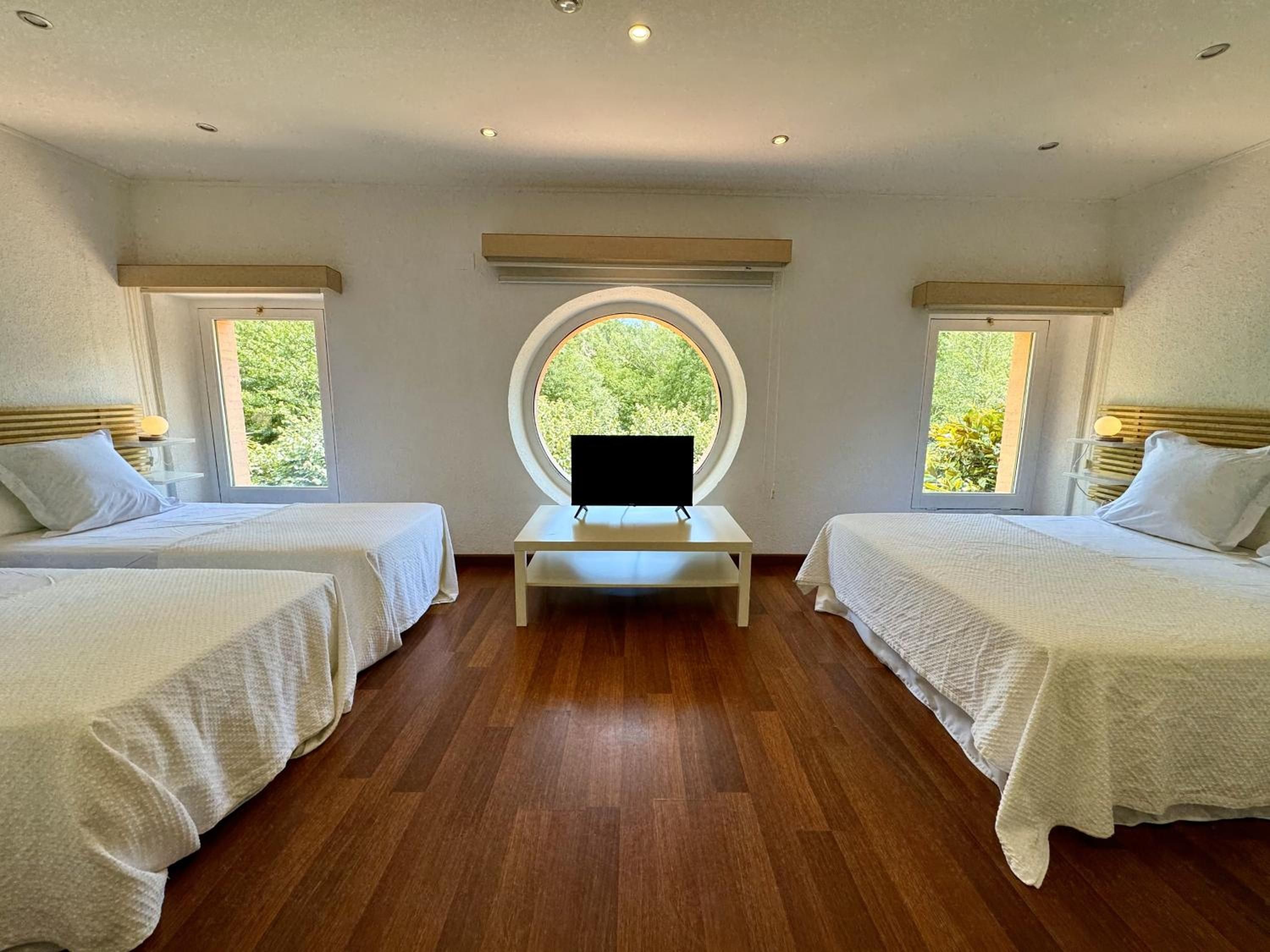 Una habitación con dos camas, una mesa central, televisión y ventanas con vistas a zonas verdes.