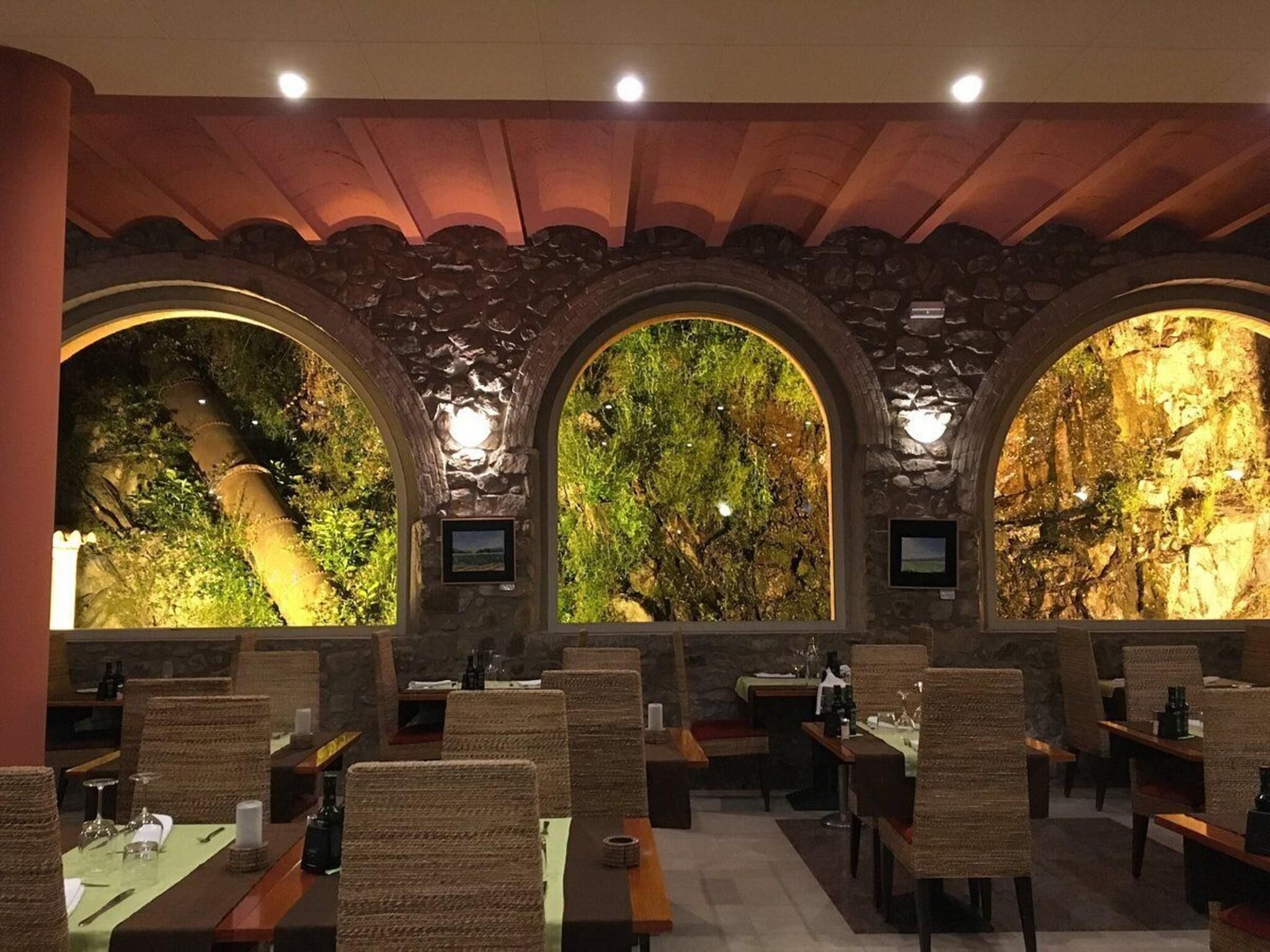 Un ristorante con interni rustici, caratterizzato da pareti in pietra e finestre ad arco che si affacciano su un paesaggio roccioso. I tavoli sono apparecchiati con sedie e l'ambiente è illuminato da luci a soffitto.