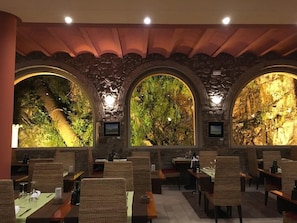 Un restaurant au décor rustique, avec des murs en pierre et des fenêtres en arc d’où l’on peut admirer un paysage rocheux. Les tables sont disposées avec des chaises, et l’espace est éclairé par des luminaires au plafond.
