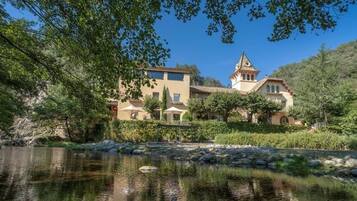 A large, multi-story building with a prominent tower, situated beside a calm body of water, surrounded by lush greenery.