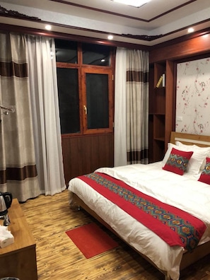 Habitación de hotel con cama de madera, ropa de cama blanca, un plaid rojo, un escritorio de madera y una ventana con cortinas.