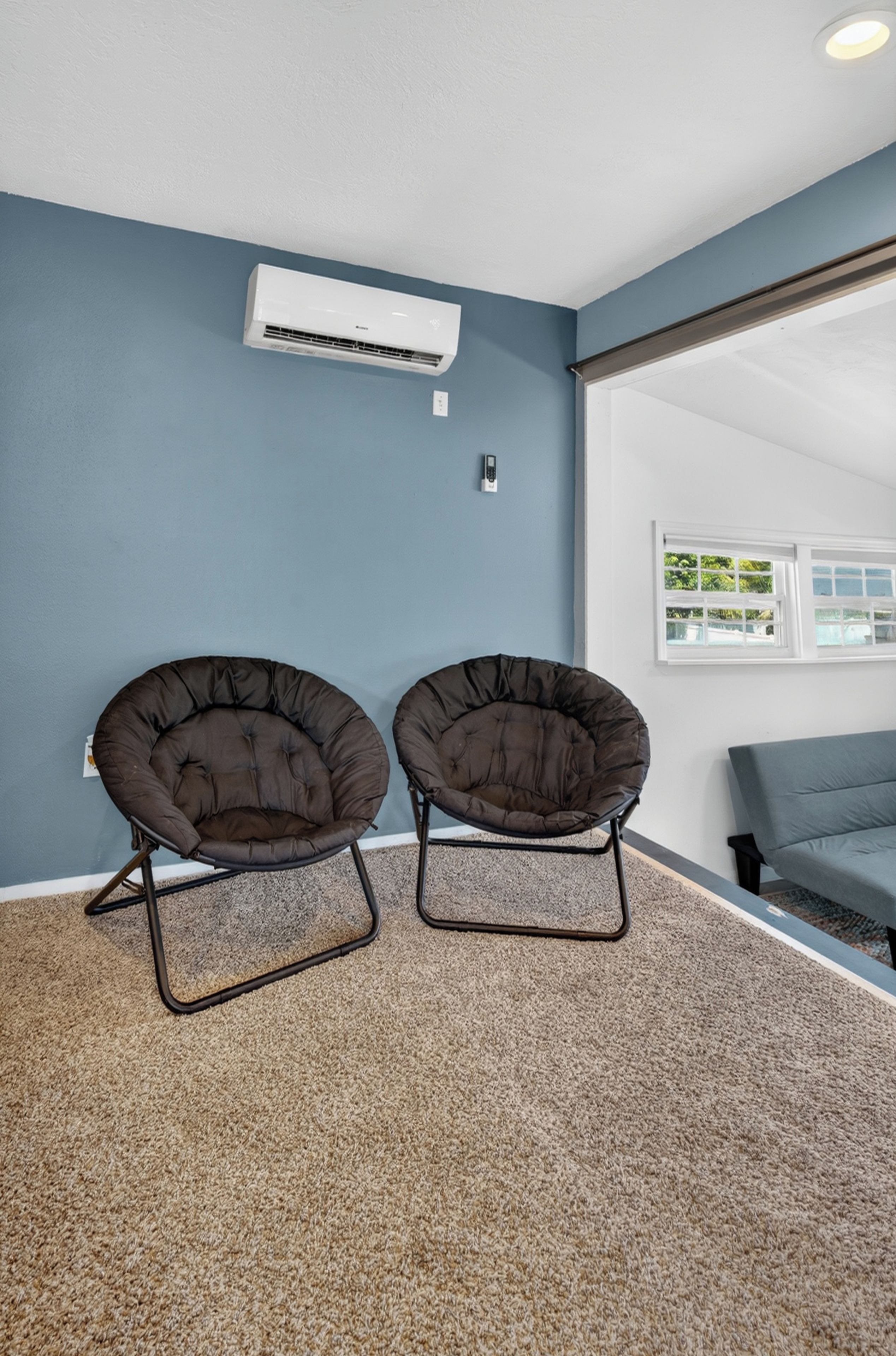 A room with two dark brown chairs, a white air conditioning unit, and a large window with a view of greenery.