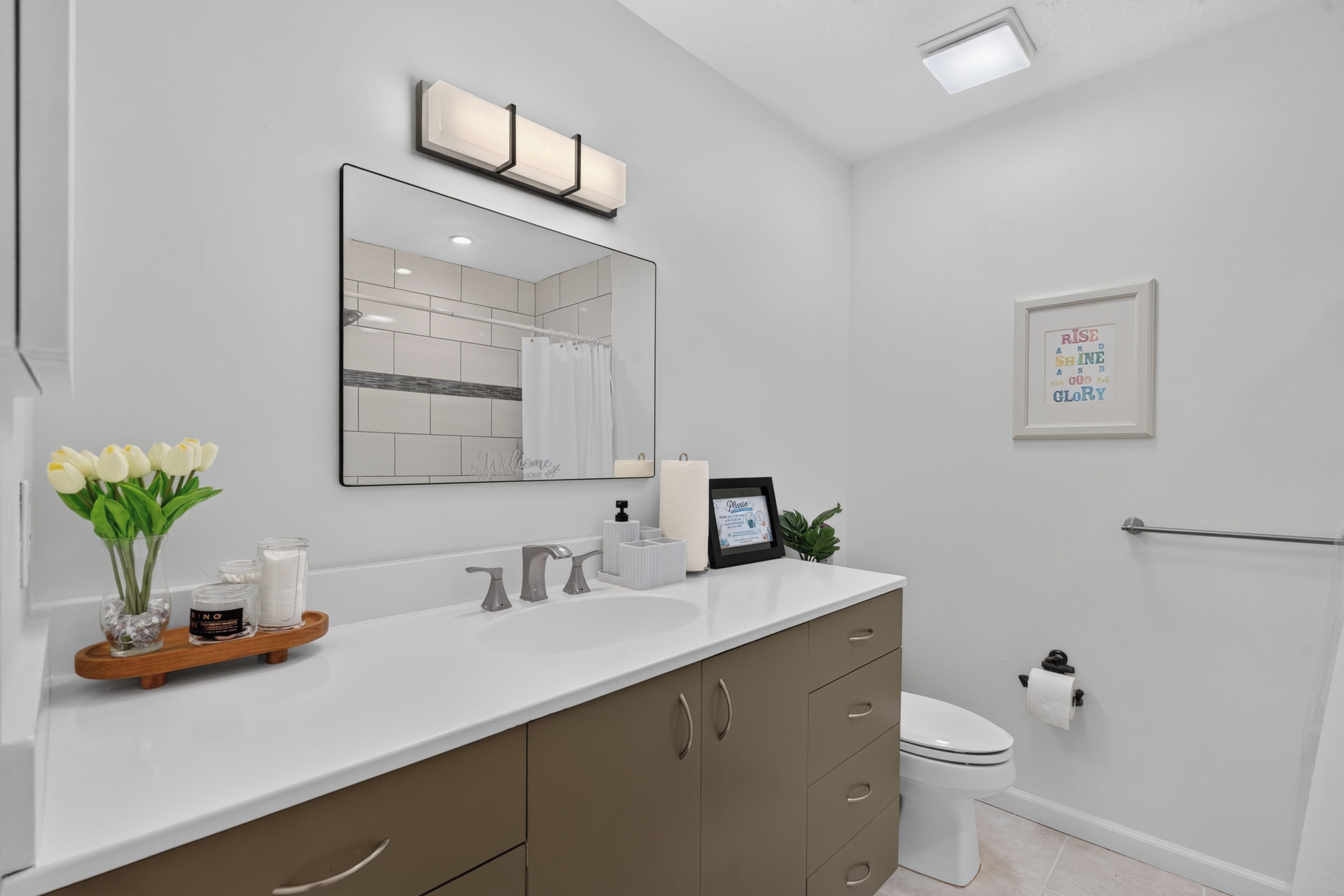 A modern bathroom with a white vanity, a large mirror, a toilet, and a framed inspirational quote on the wall.