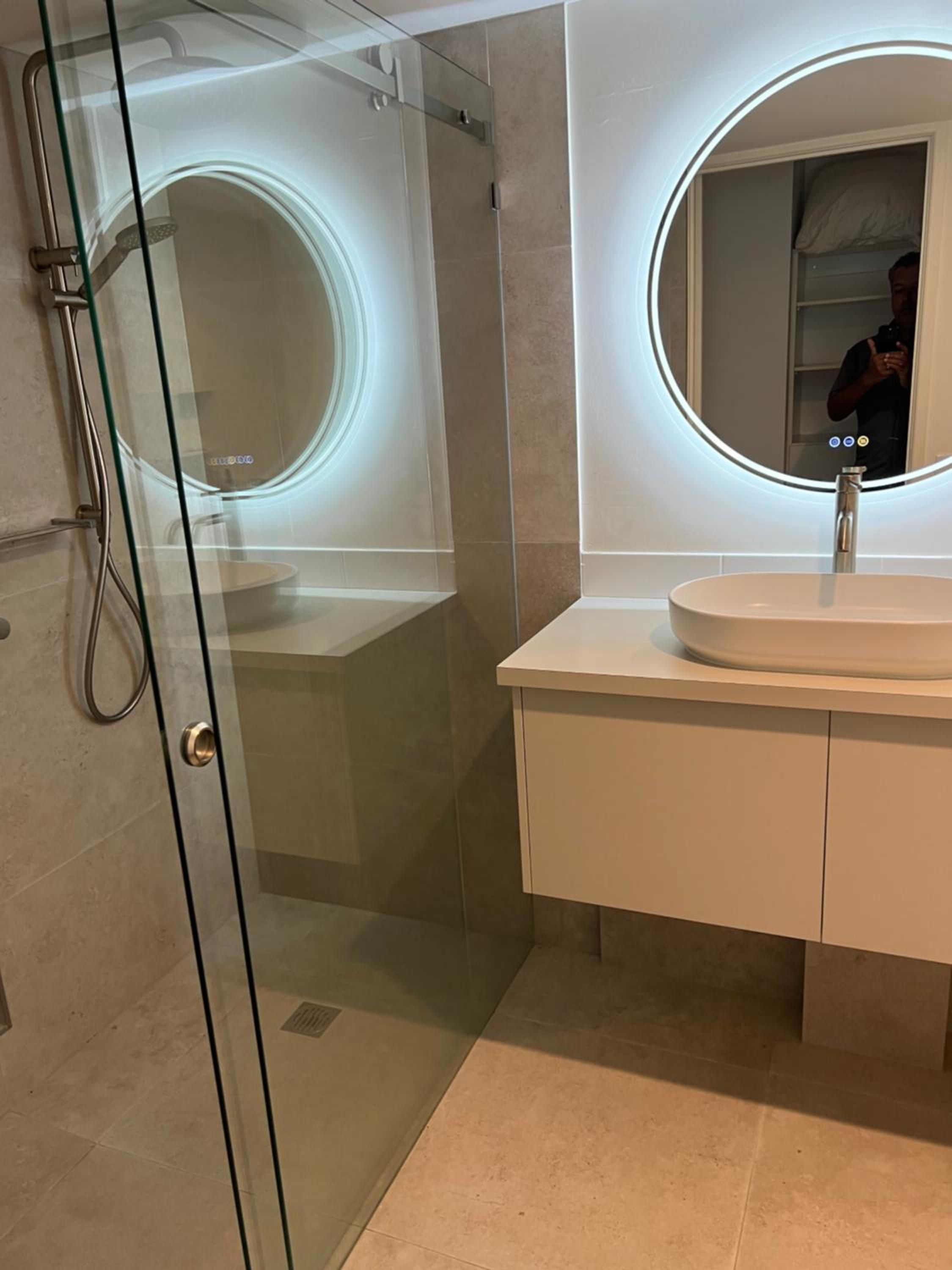 A modern bathroom with a glass-enclosed shower, a round mirror with a light, and a white sink.