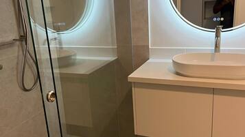 A modern bathroom with a glass-enclosed shower, a round mirror with a light, and a white sink.
