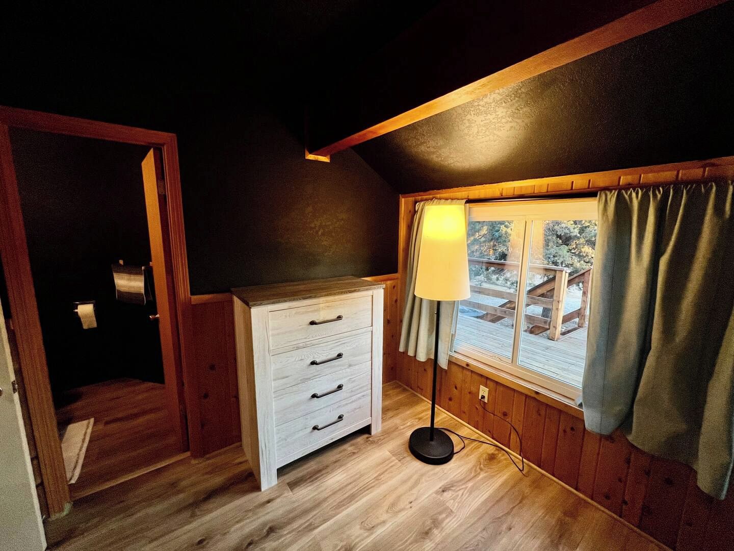 A room with a wooden floor, a white dresser with drawers, a lamp, and a window with curtains.