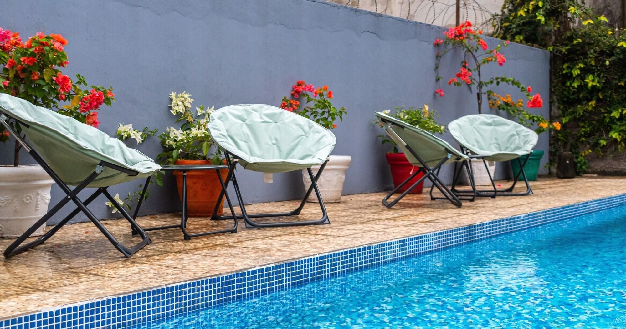 Una piscina con borde de azulejos, rodeada de sillas de descanso con fundas y plantas en macetas con flores.
