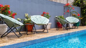 Una piscina con borde de azulejos, rodeada de sillas de descanso con fundas y plantas en macetas con flores.