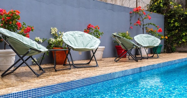 Una piscina con borde de azulejos, rodeada de sillas de descanso con fundas y plantas en macetas con flores.