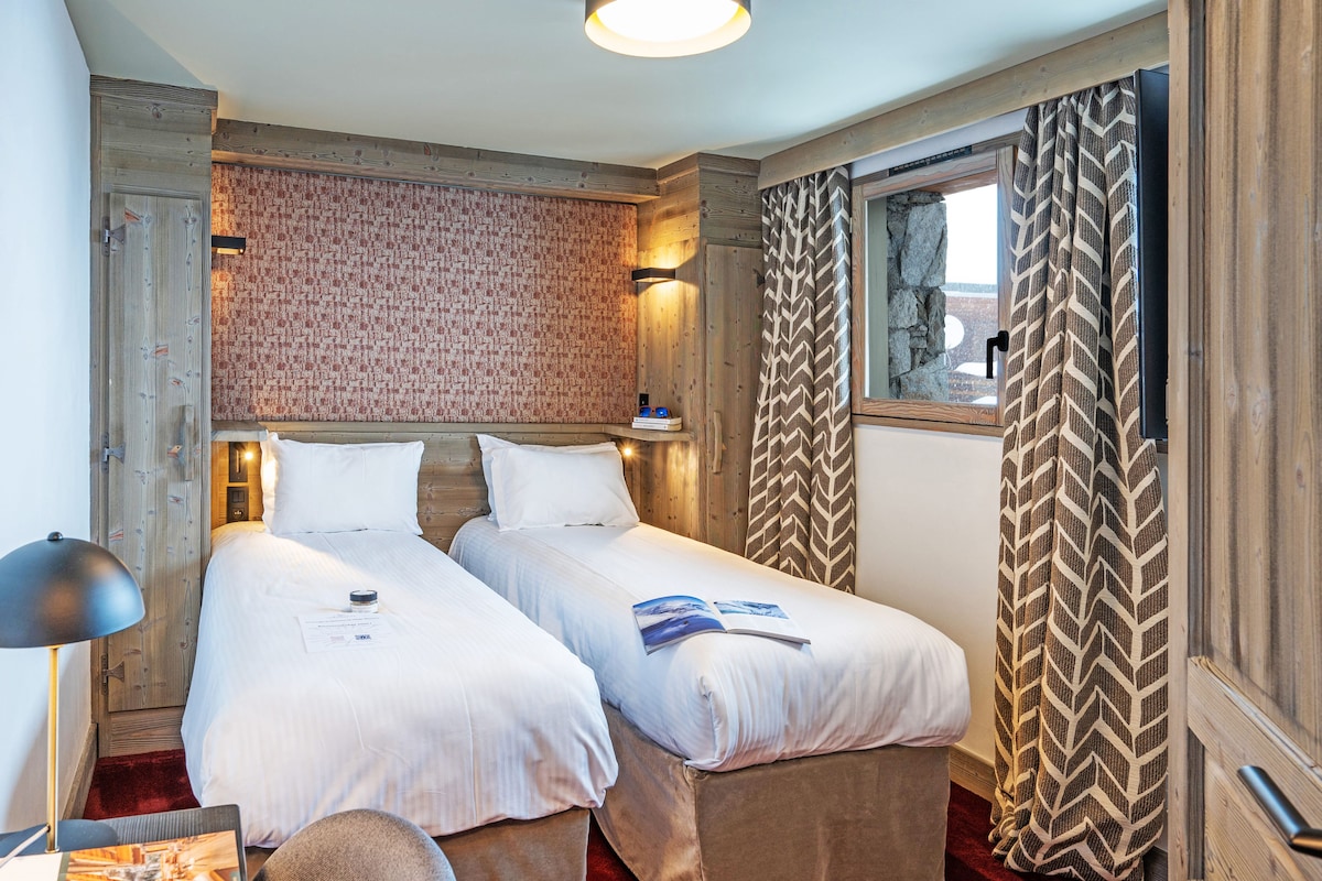 A hotel room with two single beds, a wooden headboard, a patterned wall, a window with curtains, and a nightstand with a lamp.