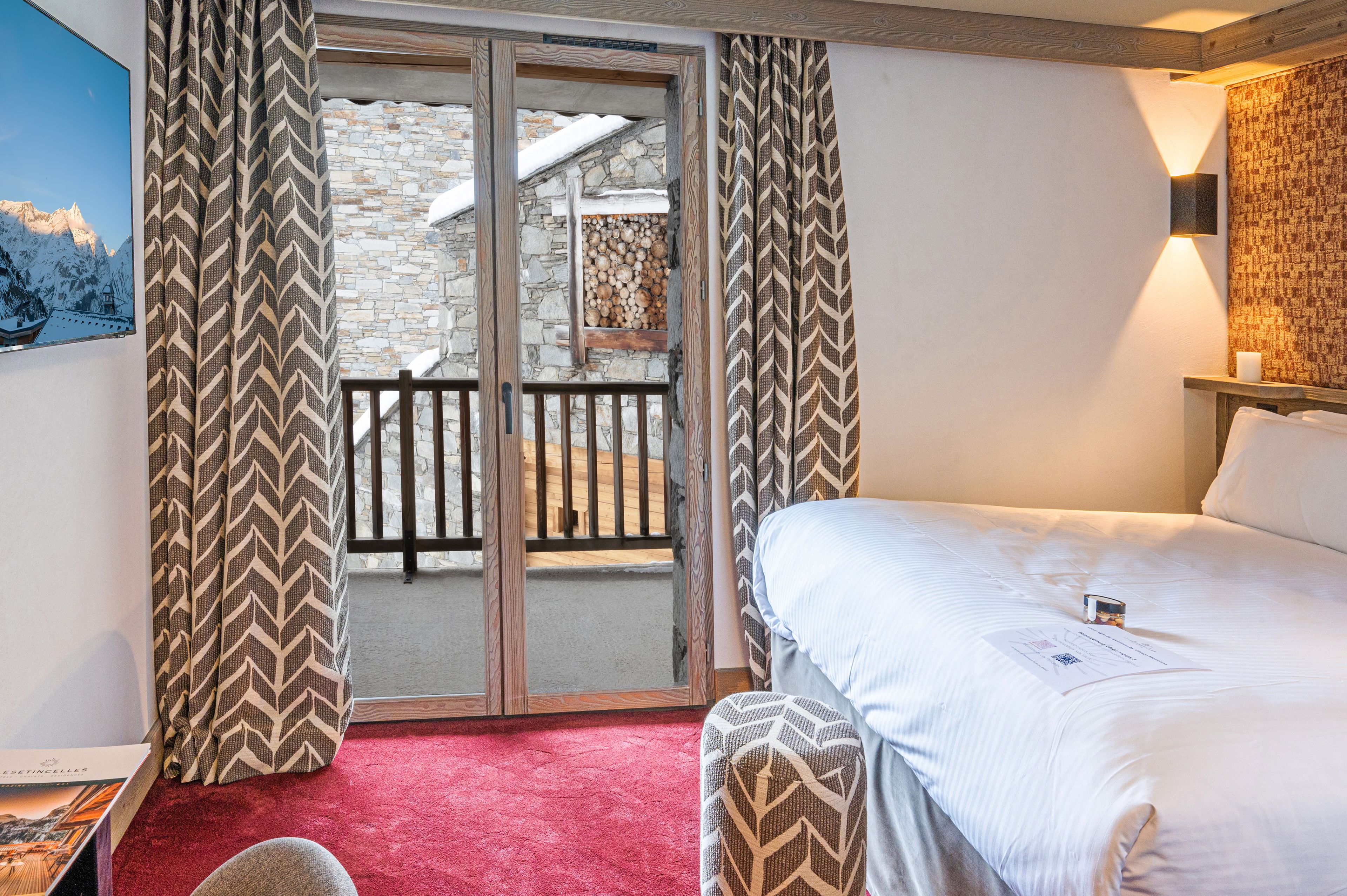 A hotel room with a bed, a red carpet, a wooden bench, a view through a glass door, and a patterned curtain.