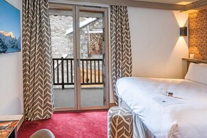 Habitación de hotel con cama, alfombra roja, banco de madera, vista por una puerta de cristal y cortina estampada.