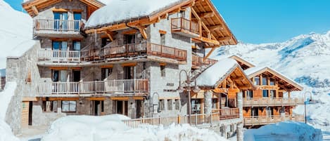 A multi-story chalet with balconies, set against a snowy mountain backdrop.
