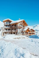 Una chaleta de varios pisos con balcones, con una imponente montaña nevada de fondo.