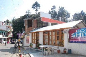 Un edificio de dos pisos con recepción y un balcón. Se observa una señalización de un hotel llamado 'HOT SHIVAY' y un número de contacto.