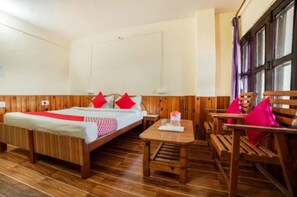 Habitación de hotel con piso de madera, una cama con almohadas rojas, una mesa de madera y un banco con cojín rosa.