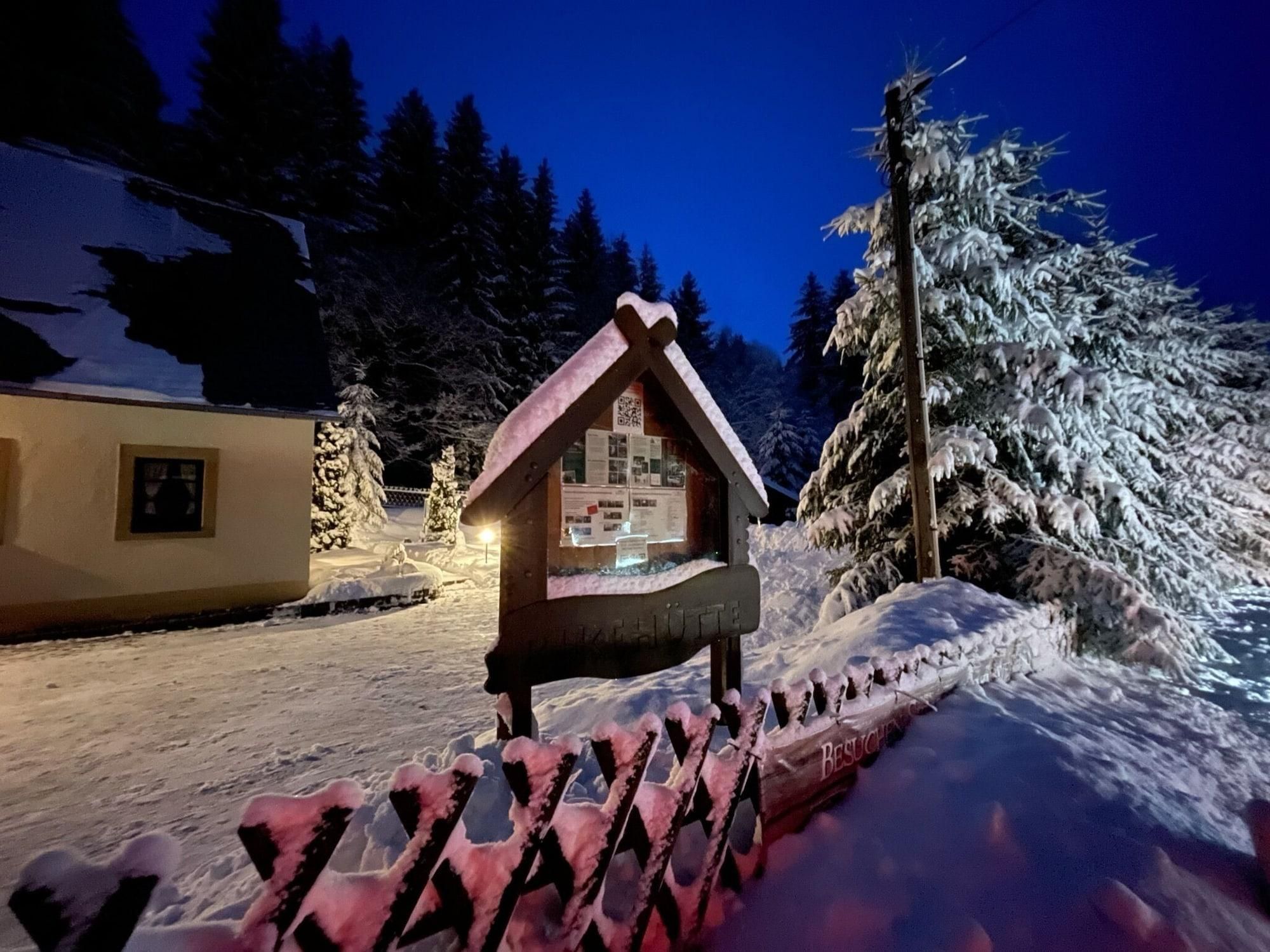 Estrutura de madeira coberta por neve com telhado inclinado, possivelmente uma cabana ou pequeno edifício, rodeada por árvores carregadas de neve e um céu noturno claro.