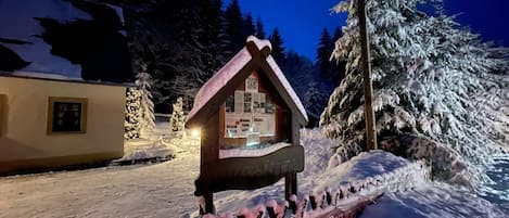 A snow-covered wooden structure with a sloped roof, possibly a cabin or a small building, surrounded by snow-laden trees and a clear night sky.
