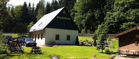 Una cabaña con techo verde identificada como 'REHNHAUS', rodeada de césped con mobiliario exterior y bicicletas.
