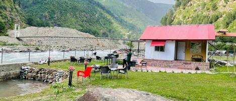 A riverside restaurant with outdoor seating, a stone retaining wall, and a mountain backdrop.