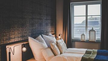 Ein Bett mit weißer Bettwäsche, ein Kopfteil mit strukturierter Gestaltung, eine Nachttischlampe und ein Fenster mit Blick auf Gebäude und eine Laterne im Fensterbrett.