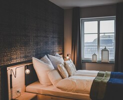 Una cama con ropa blanca, un cabecero de diseño texturizado, una lámpara de noche y una ventana con vistas a edificios y un farol en el alféizar.