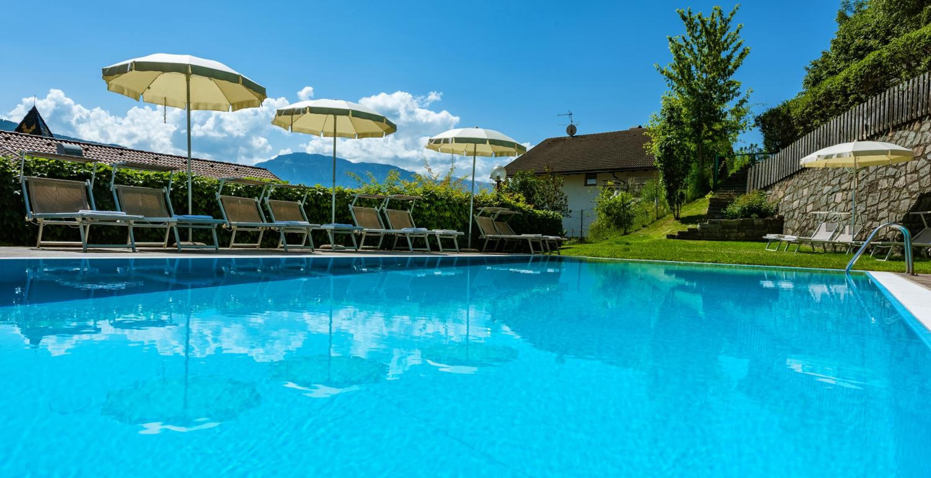 Una piscina con sillas de descanso y sombrillas, rodeada de vegetación y un muro de piedra.
