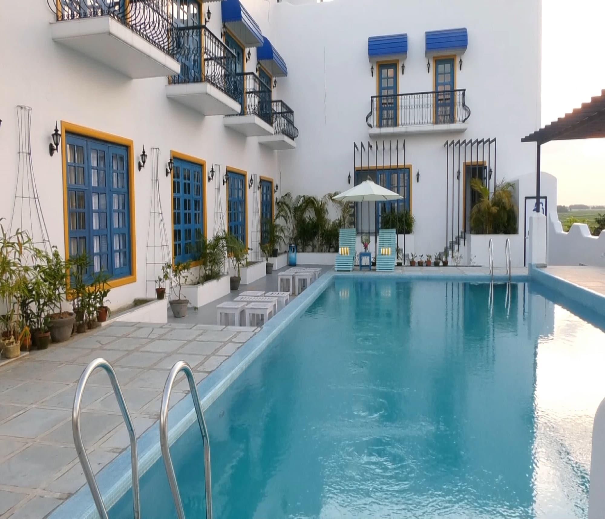 A swimming pool with a clear blue water surface, surrounded by a white building with blue doors and windows, and a small patio area with potted plants.