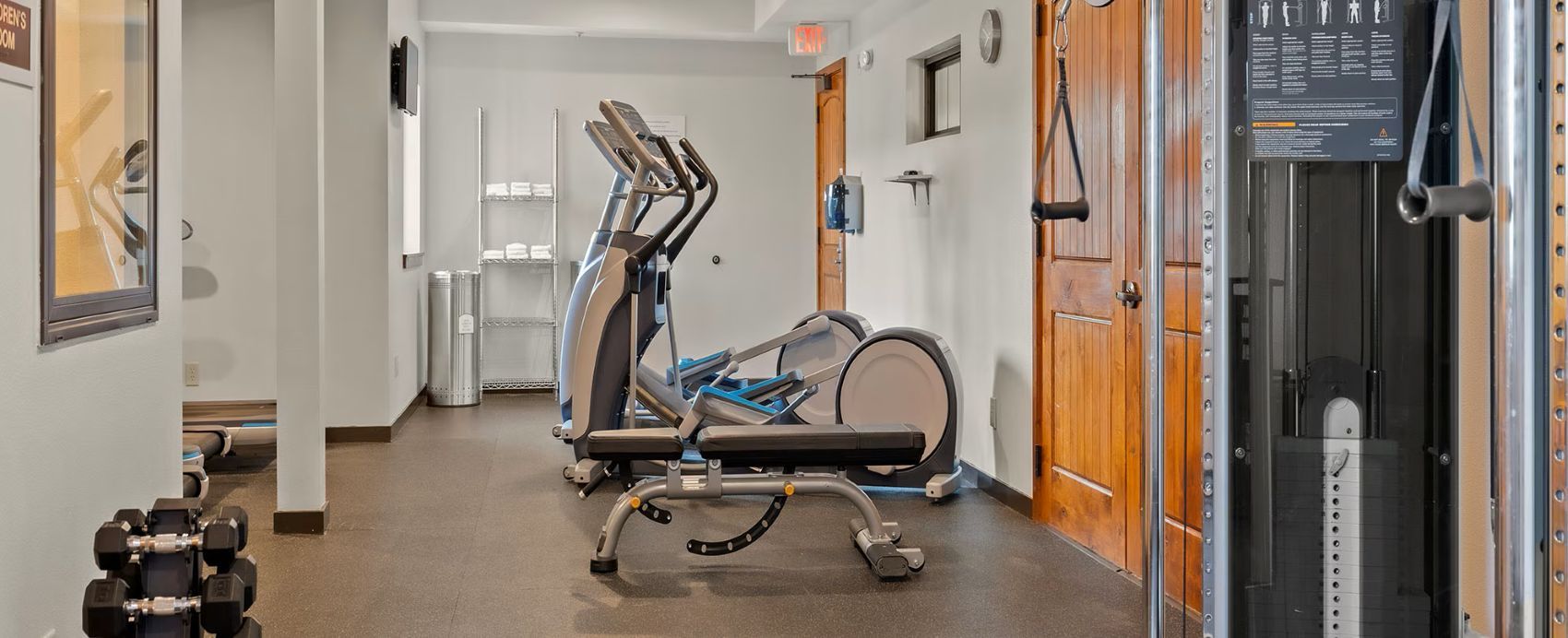A gym with exercise equipment, including a treadmill and weights, in a hallway with doors and a trash bin.