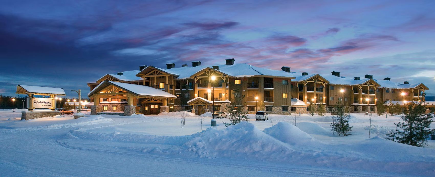 A multi-story lodge with a snow-covered roof, large windows, and a well-lit exterior.