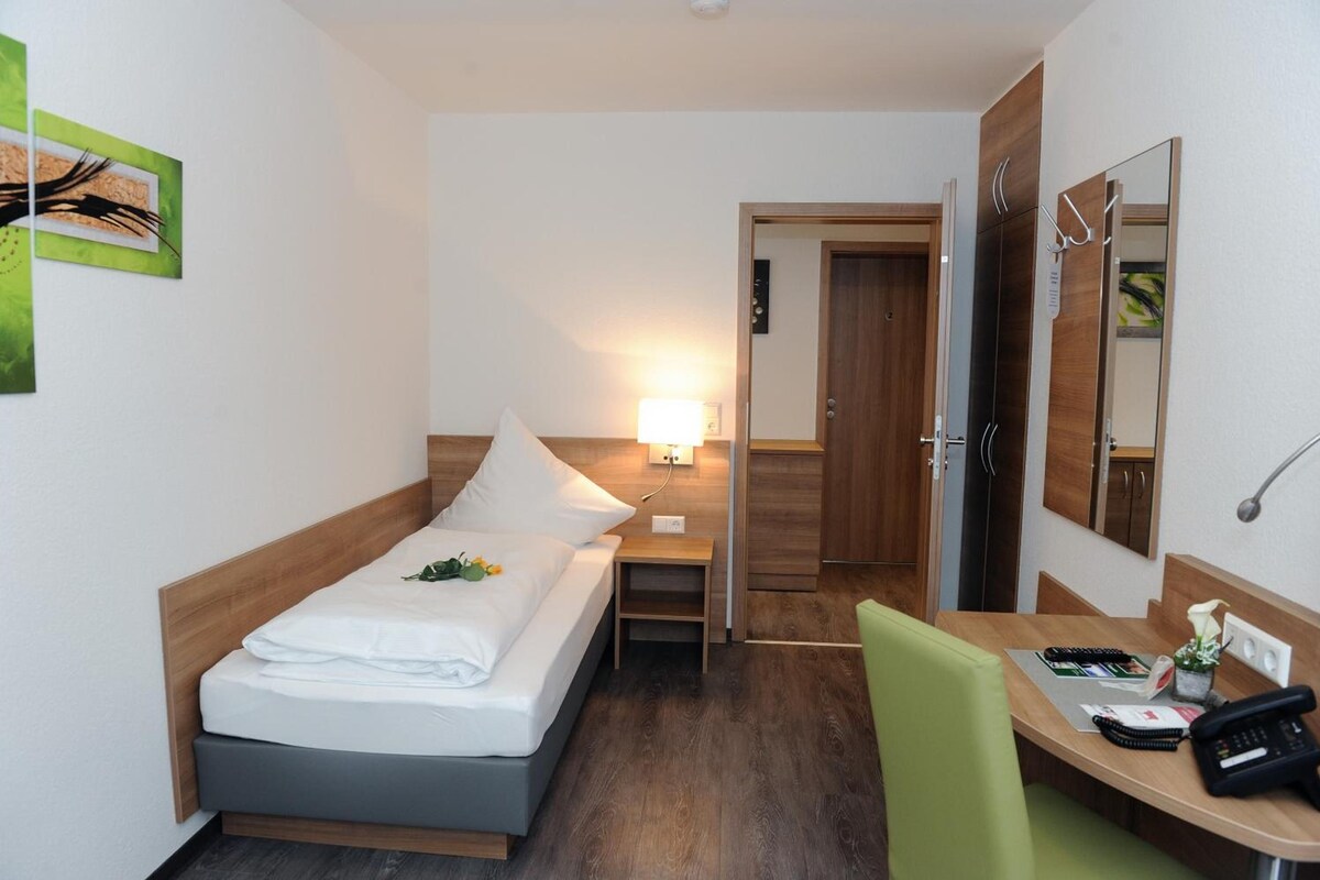 A hotel room with a bed, bedside table, lamp, chair, desk with a phone, and a mirror.