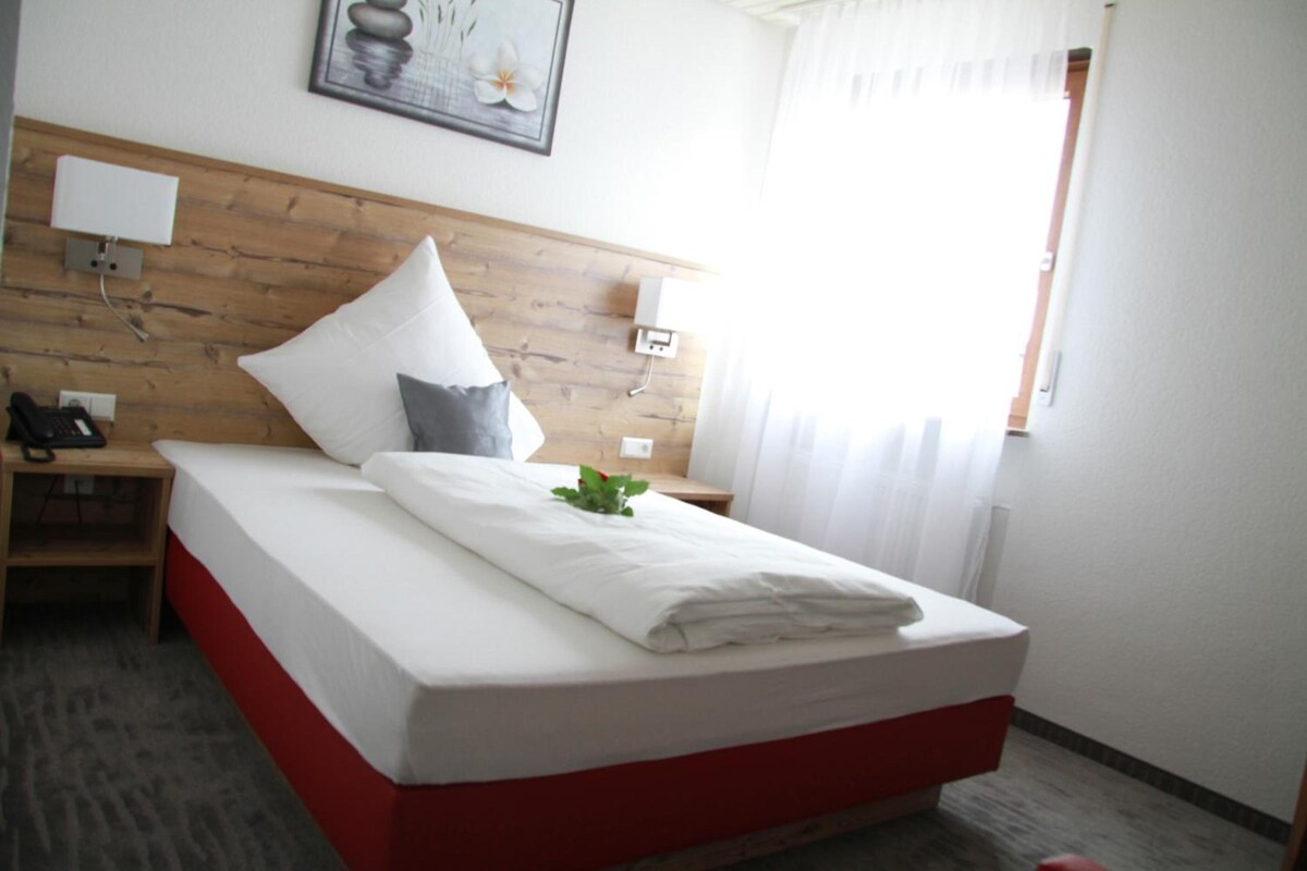 A hotel room with a wooden headboard, a bed with white bedding, a bedside table with a lamp, and a window with curtains.