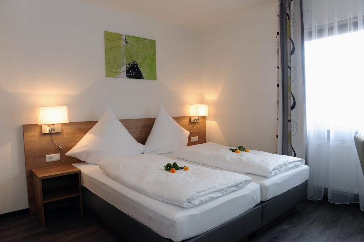 A hotel room with two beds, each with white linens and pillows, a nightstand, a wall-mounted lamp, and a painting above the beds.