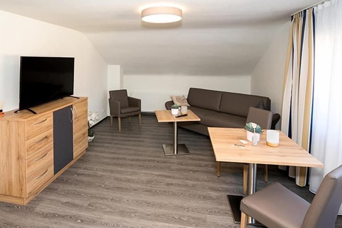 A modern living room with a flat-screen TV, a wooden cabinet, a sofa, a dining table with chairs, and a small coffee table.