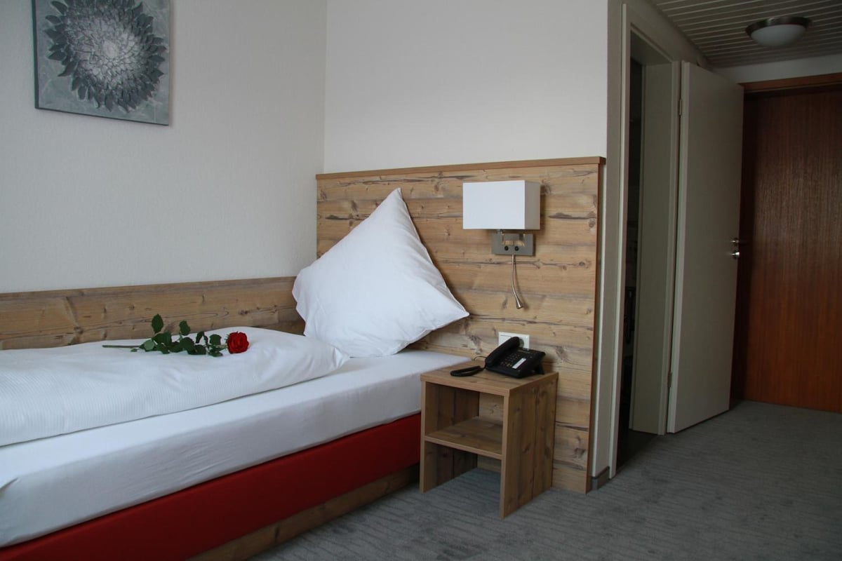 A hotel room with a wooden headboard, a single bed, a bedside table with a phone, and a door leading to another room.