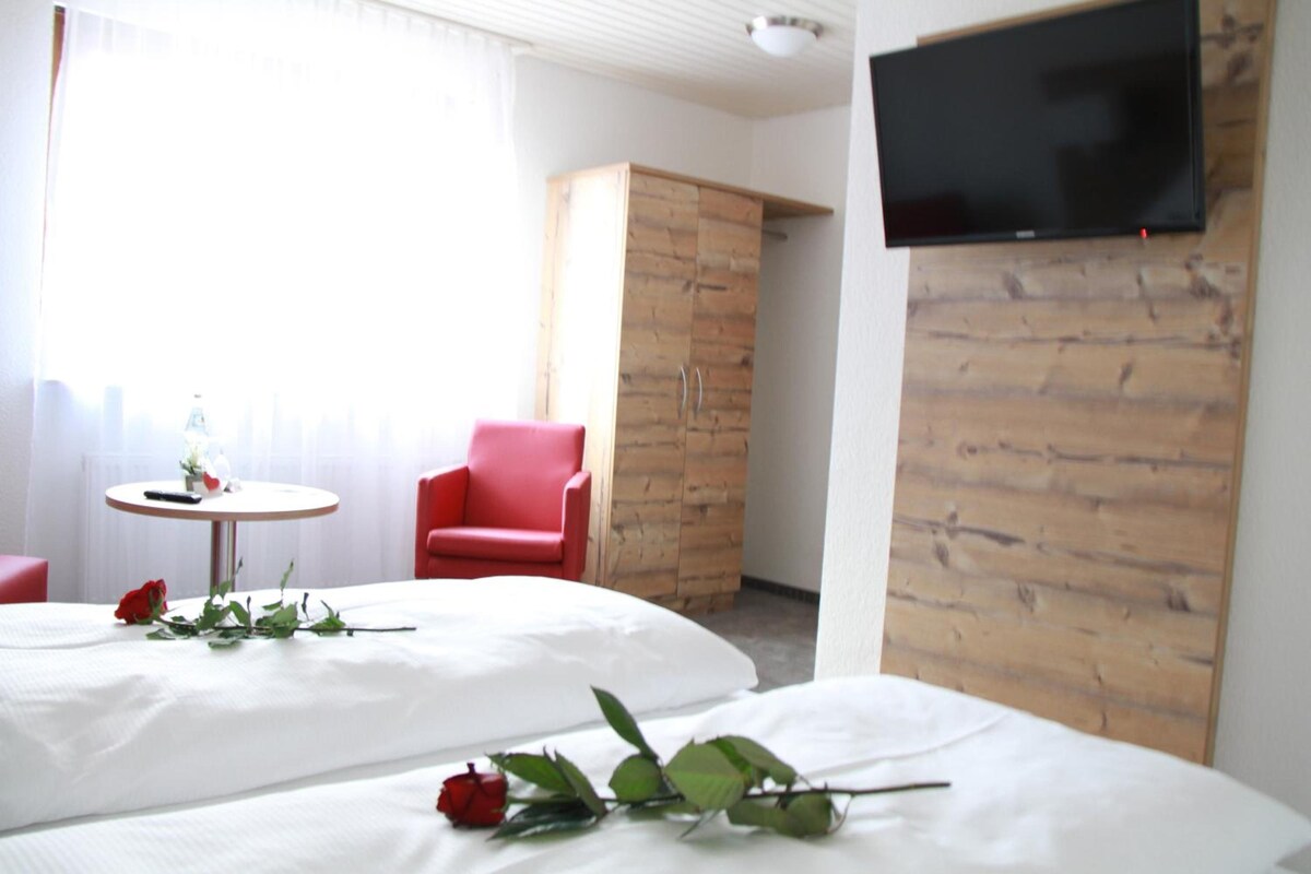 A hotel room with two beds, a red chair, a small table with a remote control, and a TV mounted on the wall.