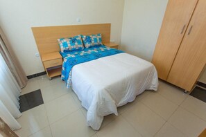 Un dormitorio con cabecera de madera, cama con sábana blanca, colcha estampada en azul y beige, mesita de noche de madera y armario.